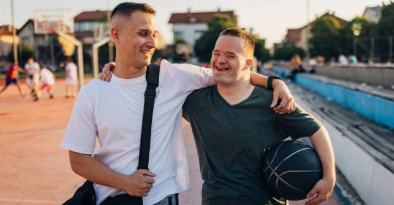 Cheerful man with Down syndrome hugs his cheerful best friend as they walk on an outdoor sports court, ready to play basketball