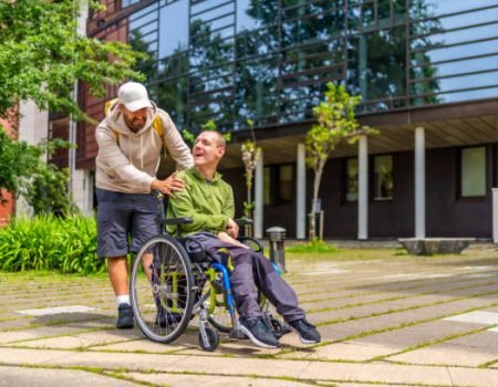 Full length photo of a caucasian adult man with special needs and friend walking along the university campus